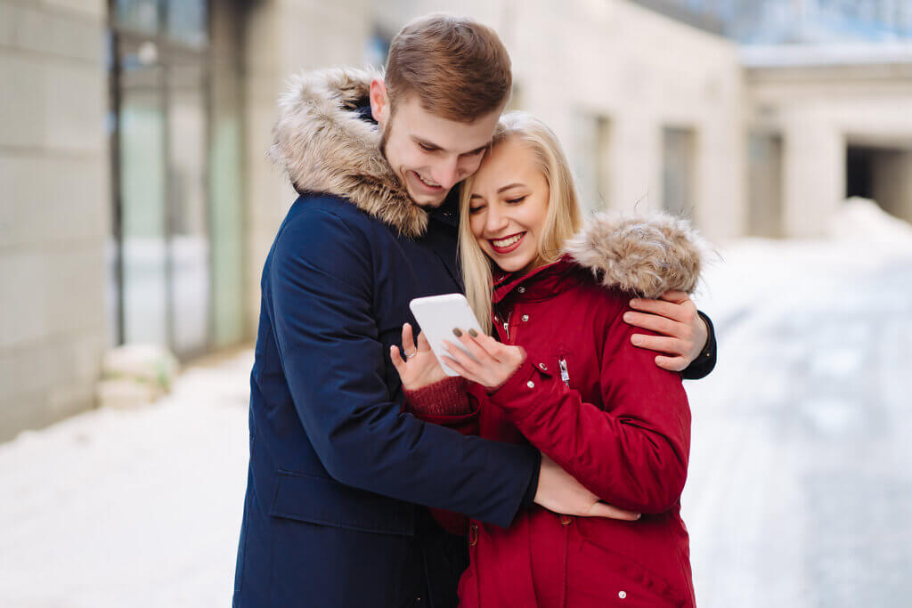 amorosa novia y novio abrazándose afuera y mirando la pantalla del teléfono móvil